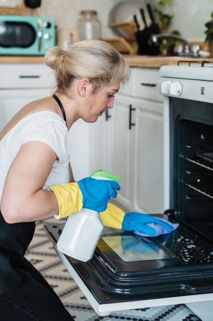 Close-up of a person's hand wearing a blue and yellow rubber glove, using a blue cloth to clean the glass surface of a black oven door. The oven is open, revealing the interior with metal racks. The s