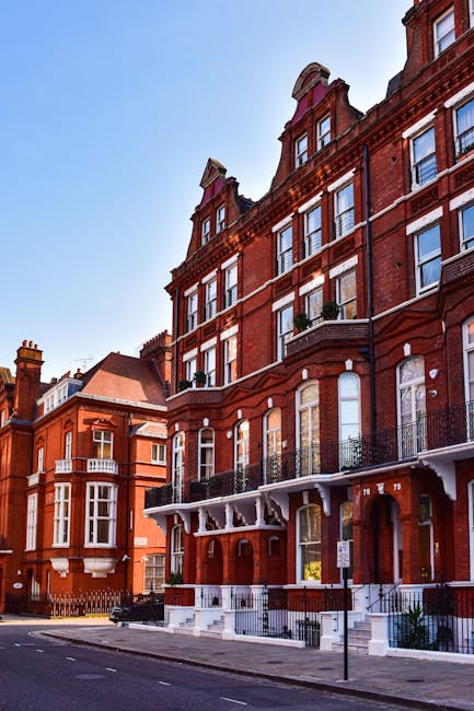 The image shows a row of Victorian-style brick residential buildings with large windows and decorative architectural details, situated along a quiet street under a clear blue sky. The buildings featur