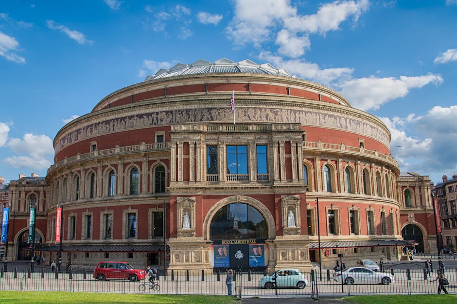 A wide view of the Royal Albert Hall in South Kensington, London, showcasing its historic circular architecture with a domed roof and ornate stonework. The building's exterior features red brick and detailed stone accents, with multiple arched windows and decorative sculptures near the entrance. The sky above is mostly clear with a few scattered clouds. In the foreground, a busy street scene includes parked cars, bicycles, and pedestrians walking along the pavement, with a low fence separating the road from the building. This image highlights a London landmark in a vibrant, urban setting, captured in natural daylight. For professional cleaning and maintenance of such historic surfaces, visit Oven Cleaning Kensington for expert domestic cleaning services near the Royal Albert Hall.