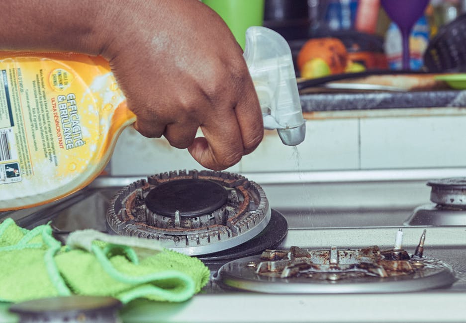 A residential kitchen scene showing a person wearing yellow and blue rubber cleaning gloves using a blue microfiber cloth to clean the surface of a white gas stove with two burners, one of which has s